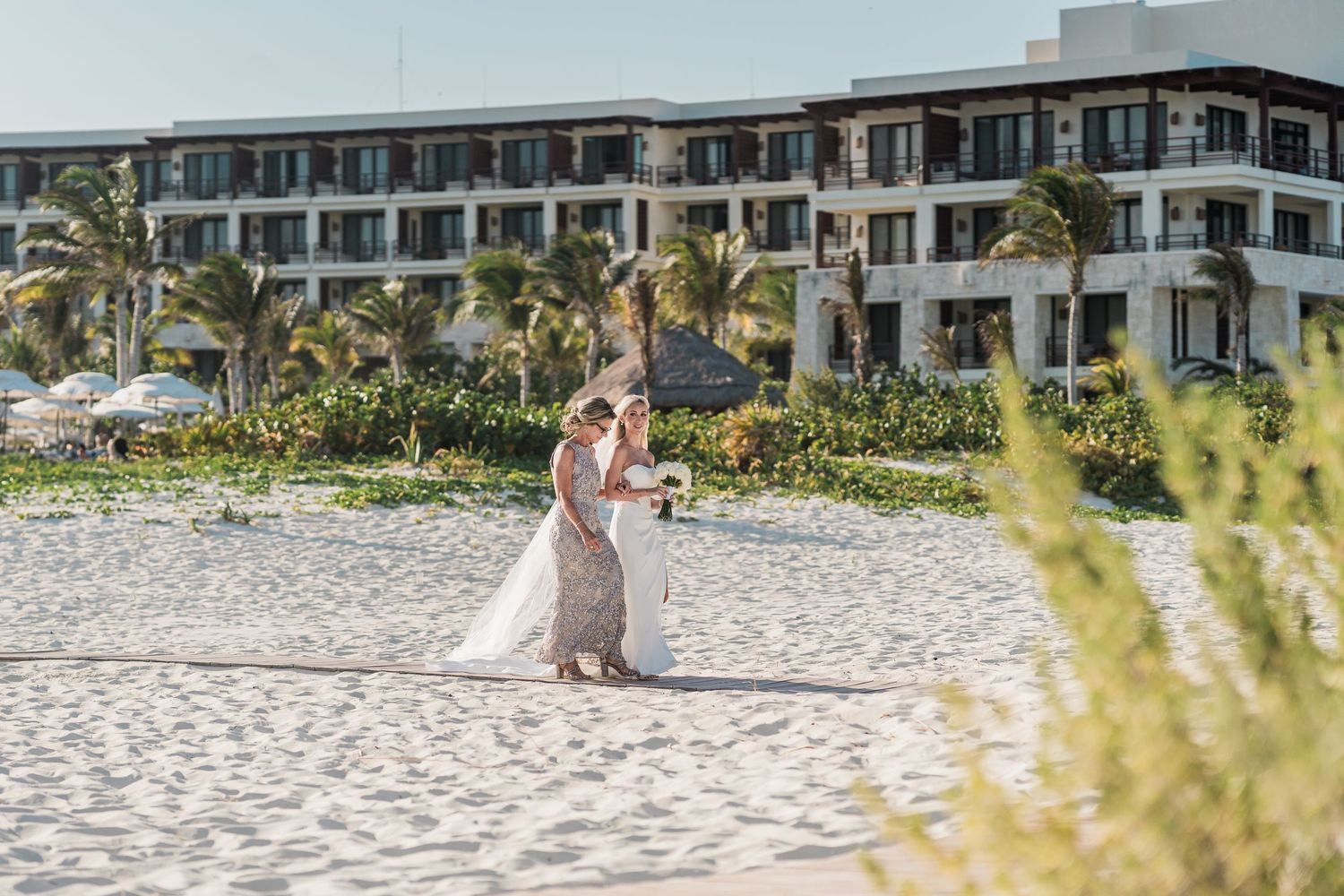 Bride in white gown walks with escort across white sand beach at tropical destination wedding.