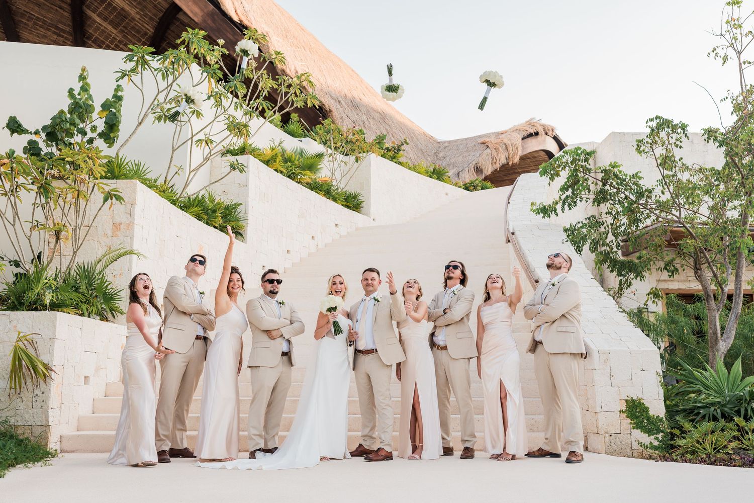 Wedding party in neutral attire poses together outdoors at a tropical resort venue with lush greenery.