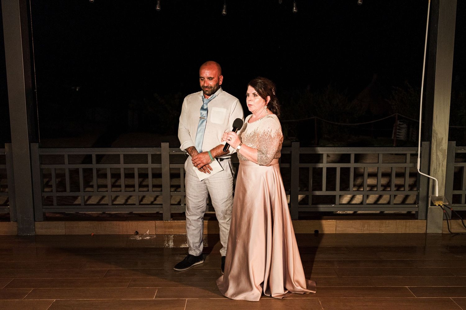 Couple in formal attire sharing a microphone while giving a speech at an outdoor evening wedding.