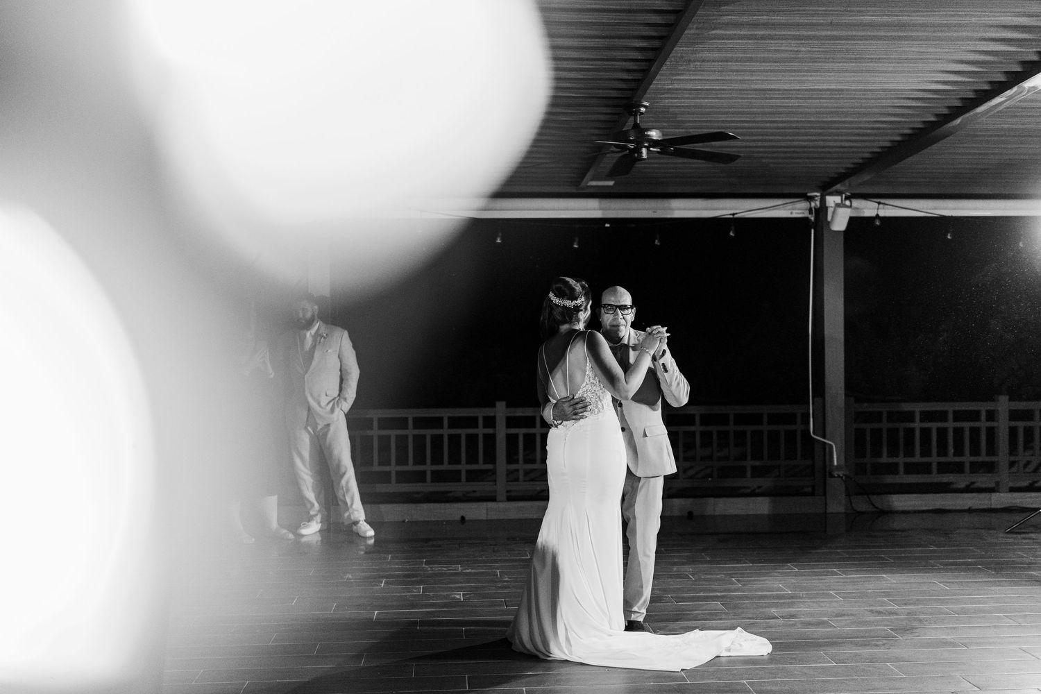 Bride and groom share their first dance in black and white wedding photo with soft bokeh foreground lights.