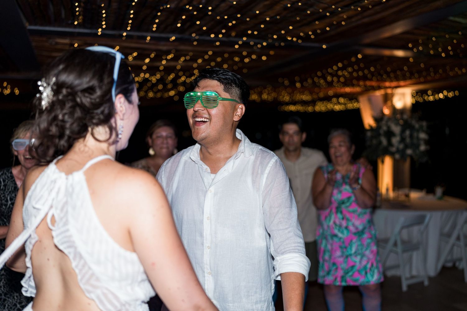 Man in white shirt wearing green sunglasses dancing with woman at wedding reception.