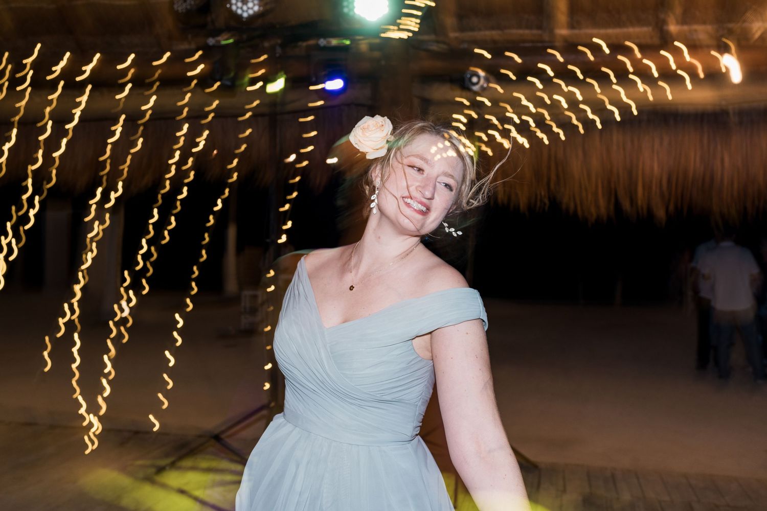 Bridesmaid in light blue dress dancing joyfully at outdoor wedding reception with bokeh string lights.