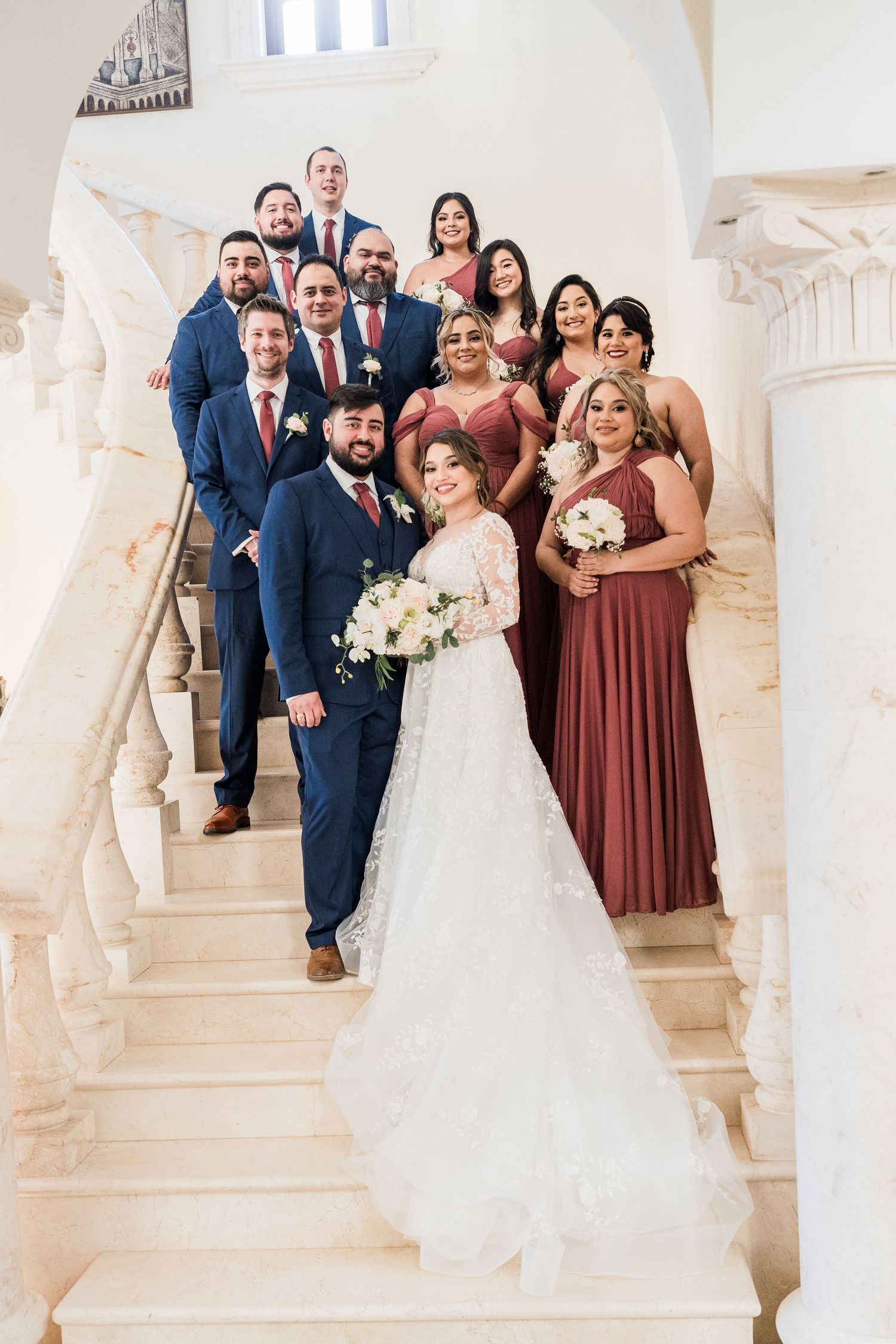 Wedding party in navy suits and dusty rose gowns posing on grand marble staircase with couple.