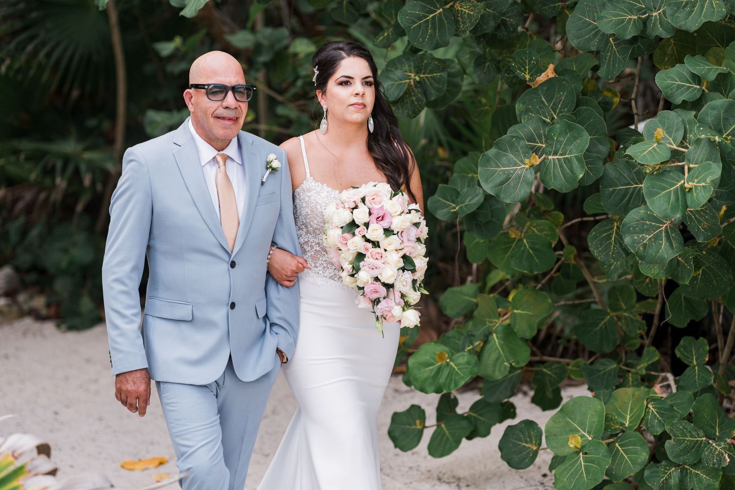 Father escorts bride in elegant white gown holding pink bouquet along tropical beach wedding path.