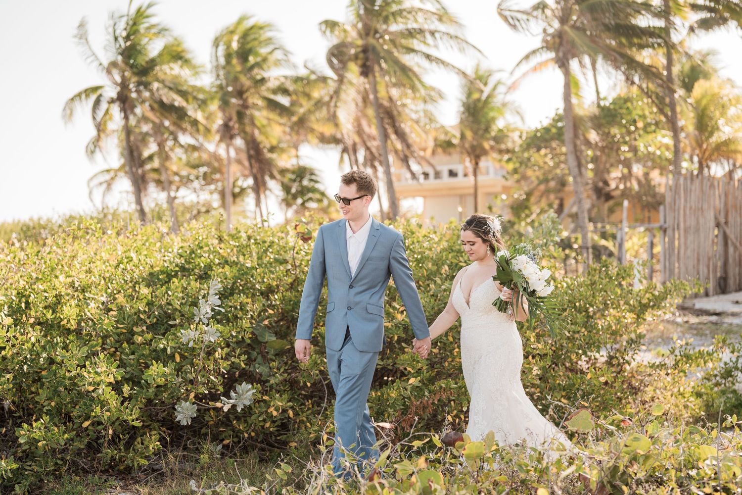 Bride and groom walk hand in hand through tropical greenery on their beach wedding day.