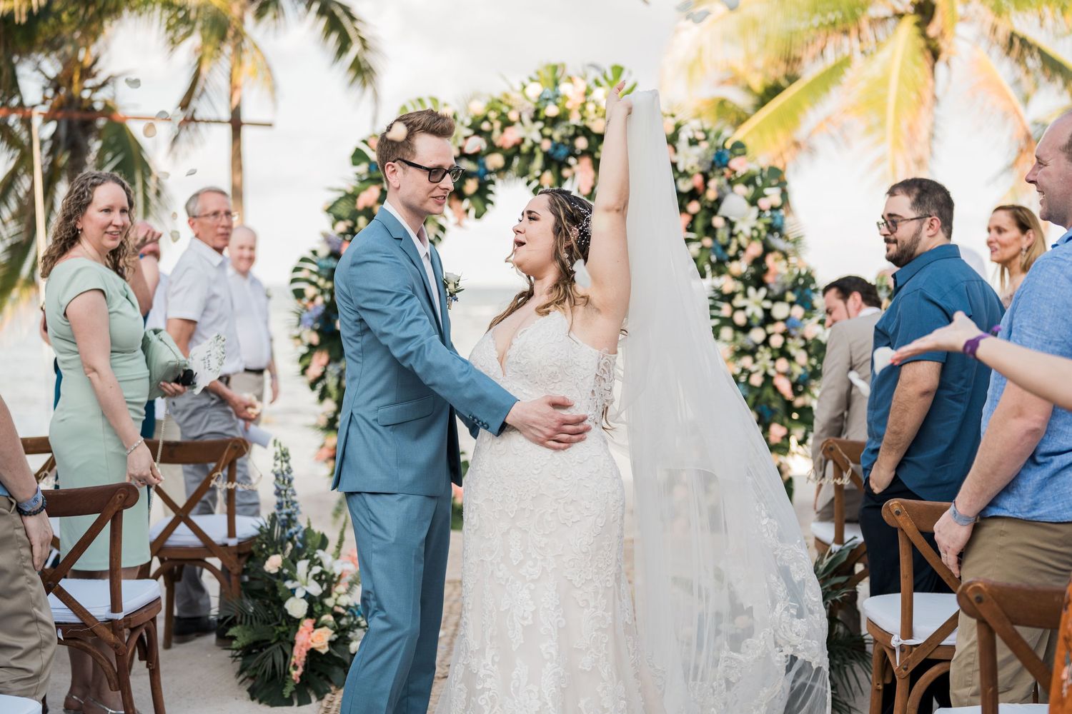 Joyful bride raises hand in celebration with groom at tropical beach wedding ceremony.