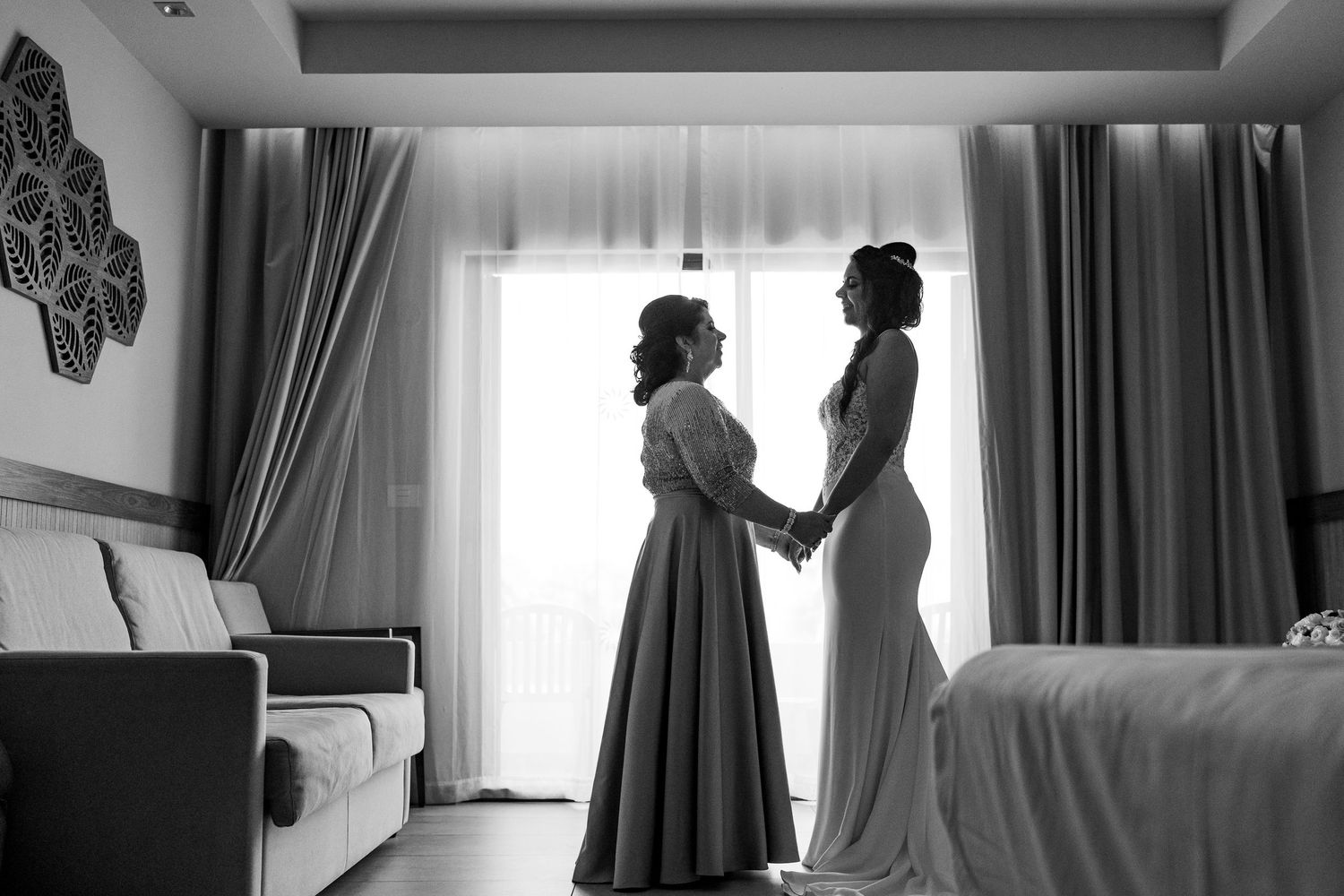 Black and white photo of bride and bridesmaid holding hands in a sunlit hotel room before the wedding.