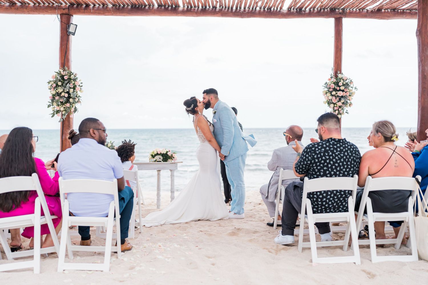 Newlyweds share their first kiss at a beautiful beachside wedding ceremony with seated guests.