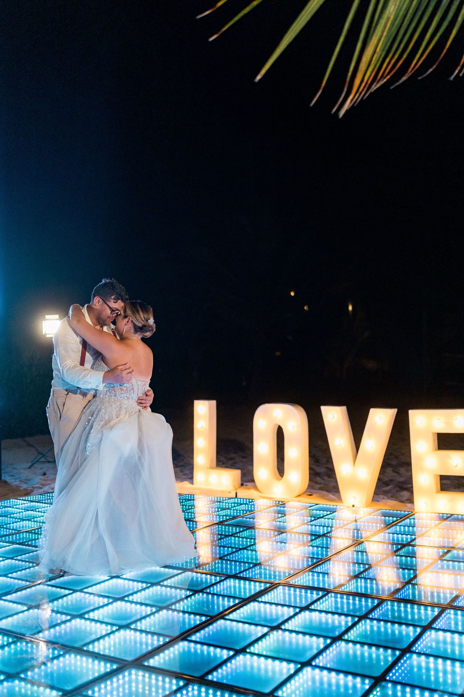 Bride and groom sharing romantic kiss on glowing LED dance floor beside illuminated LOVE sign at night.