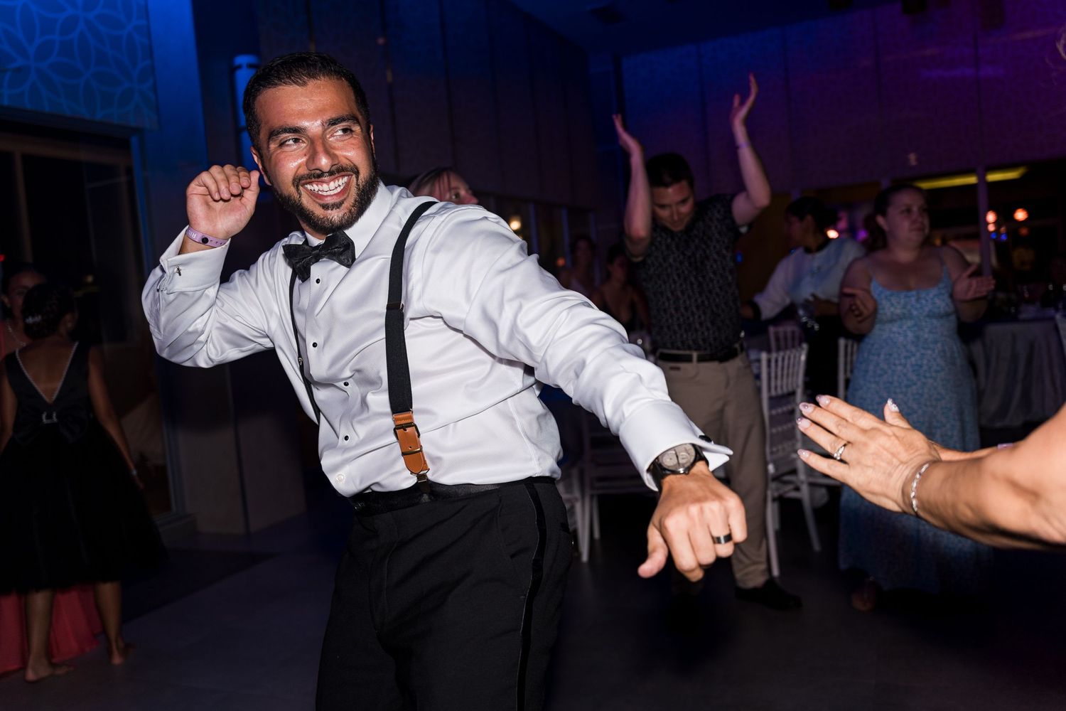 Smiling groom in suspenders and bow tie dances energetically with guests at wedding reception.