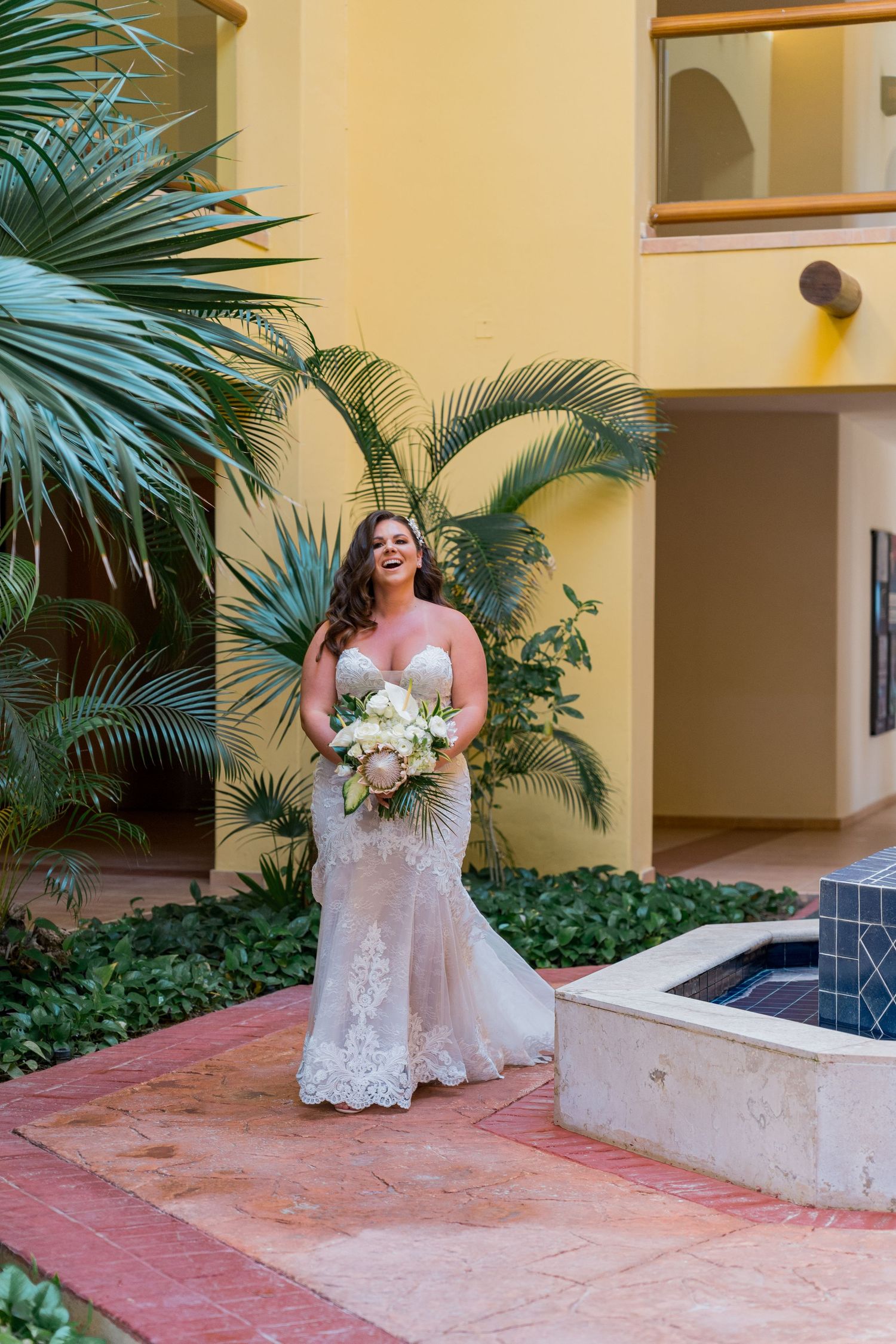 Bride in a fitted lace wedding gown holding a white bouquet standing in a tropical resort courtyard.