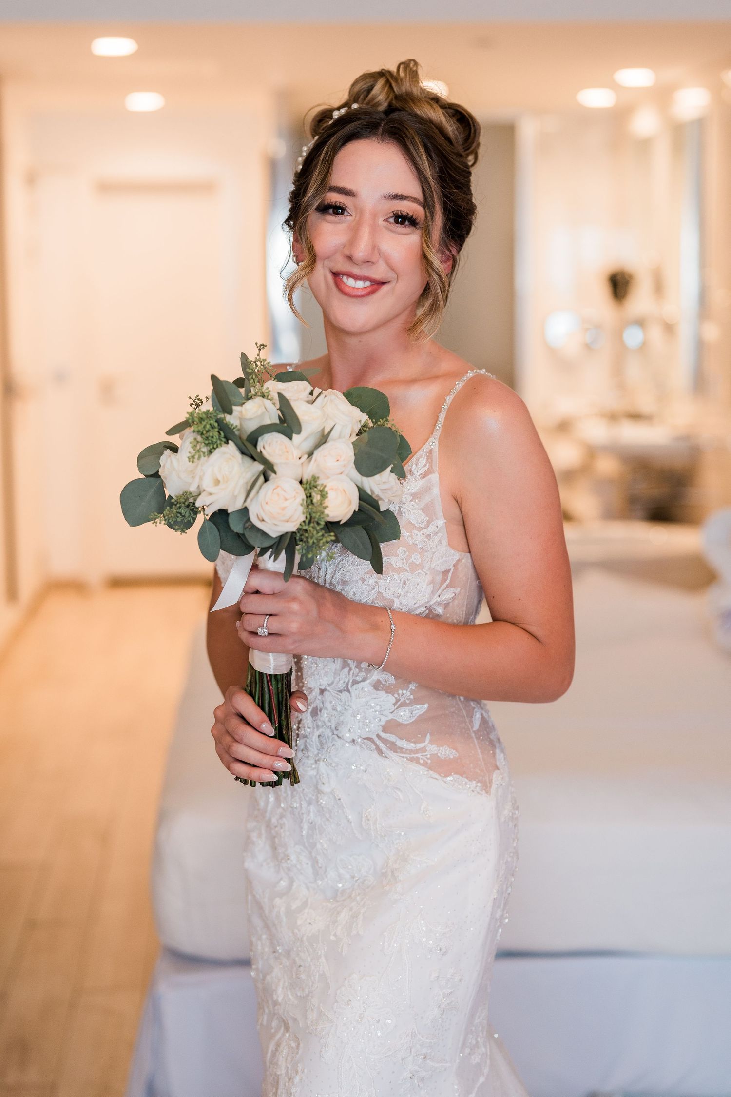 Smiling bride in lace wedding gown holding white rose bouquet indoors.