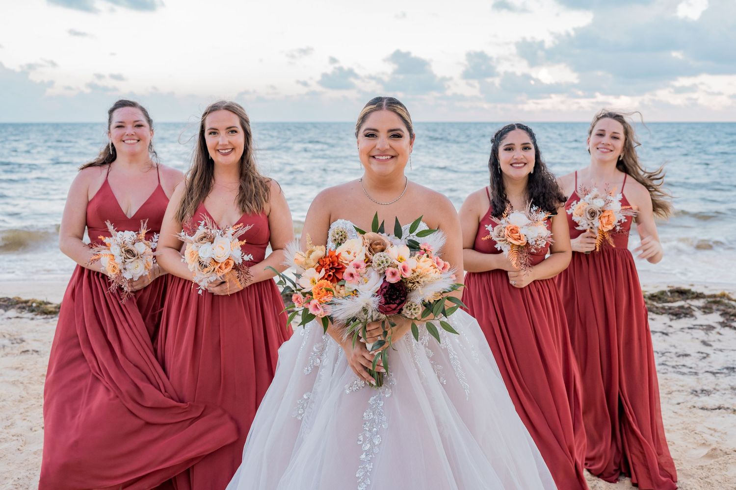 Bride with bridesmaids in burgundy dresses holding bouquets on a beach at sunset.