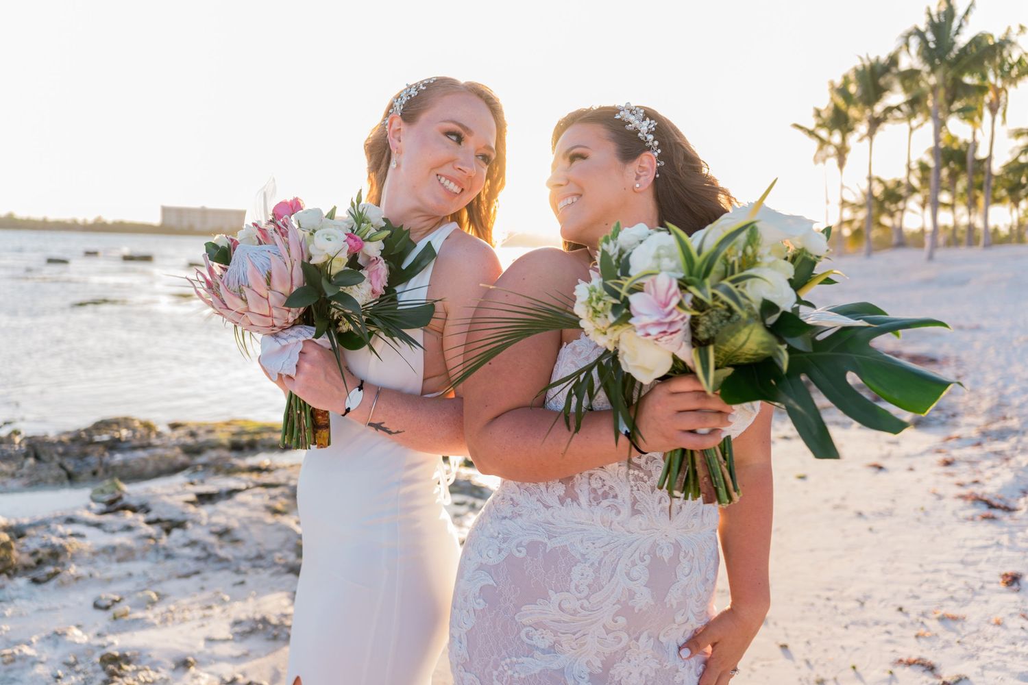 Two brides holding tropical bouquets and embracing on a sandy beach at sunset.