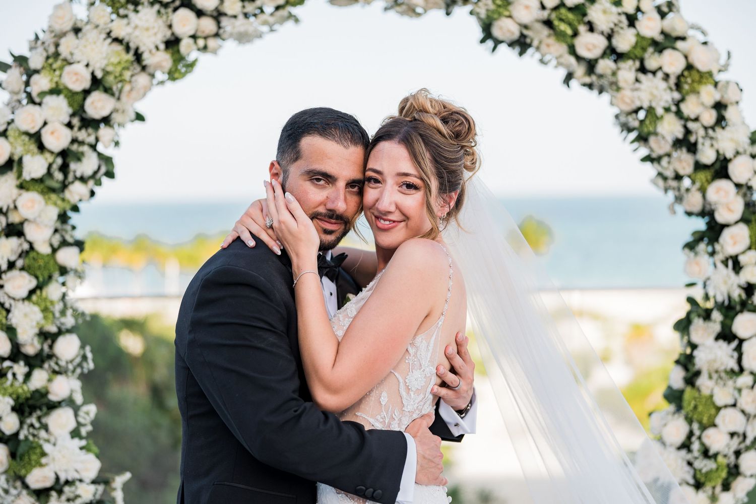 Bride and groom embrace lovingly under elegant white floral circular arch by the ocean.