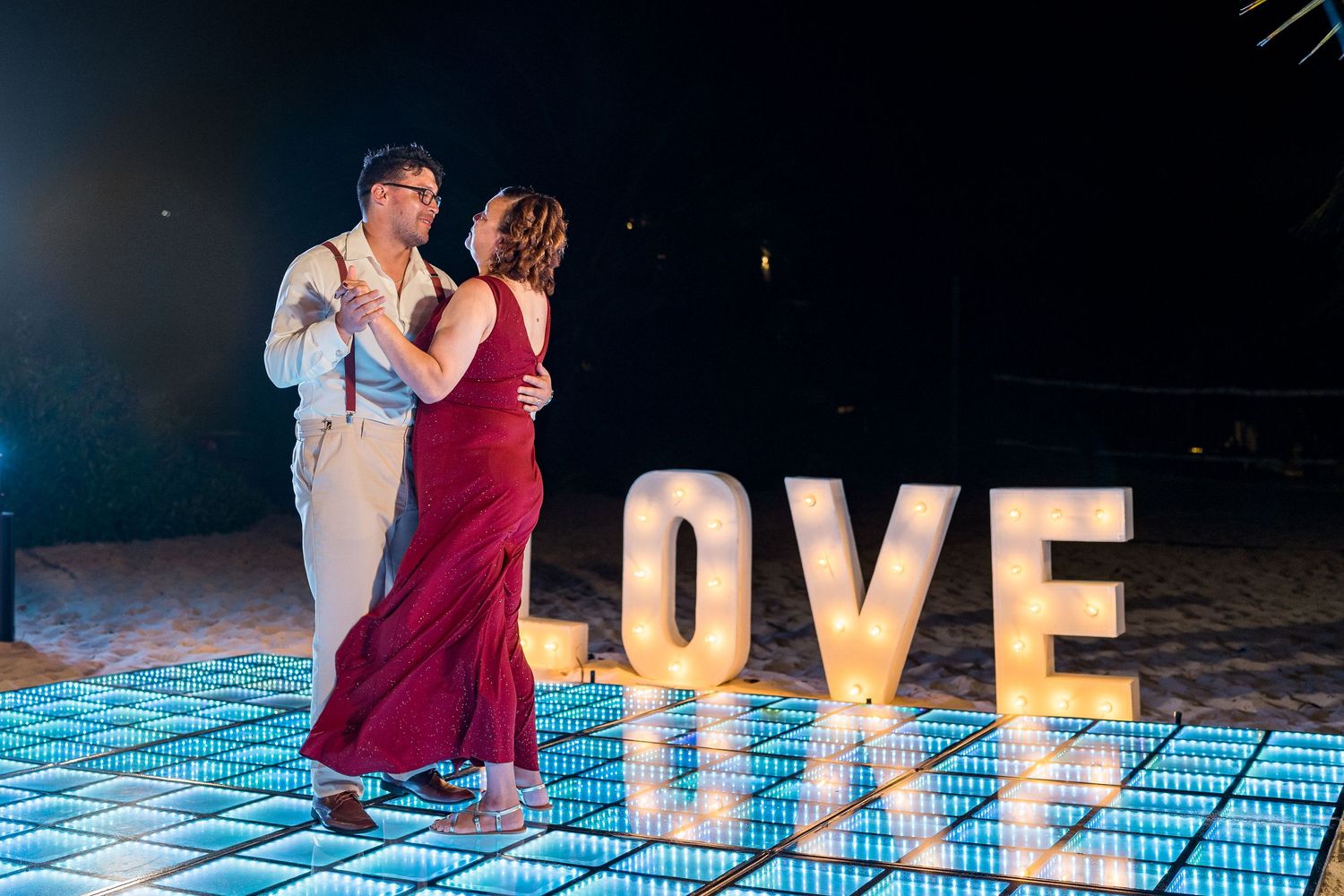 Couple dancing on LED dance floor next to glowing LOVE marquee letters at beach wedding.