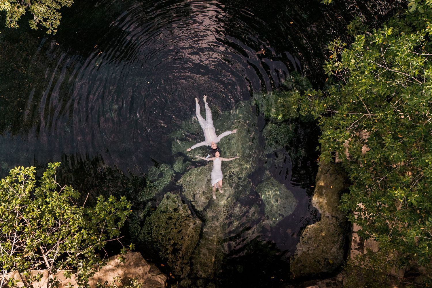 Aerial view of a person floating in a dark natural swimming hole surrounded by rocks and lush green trees.