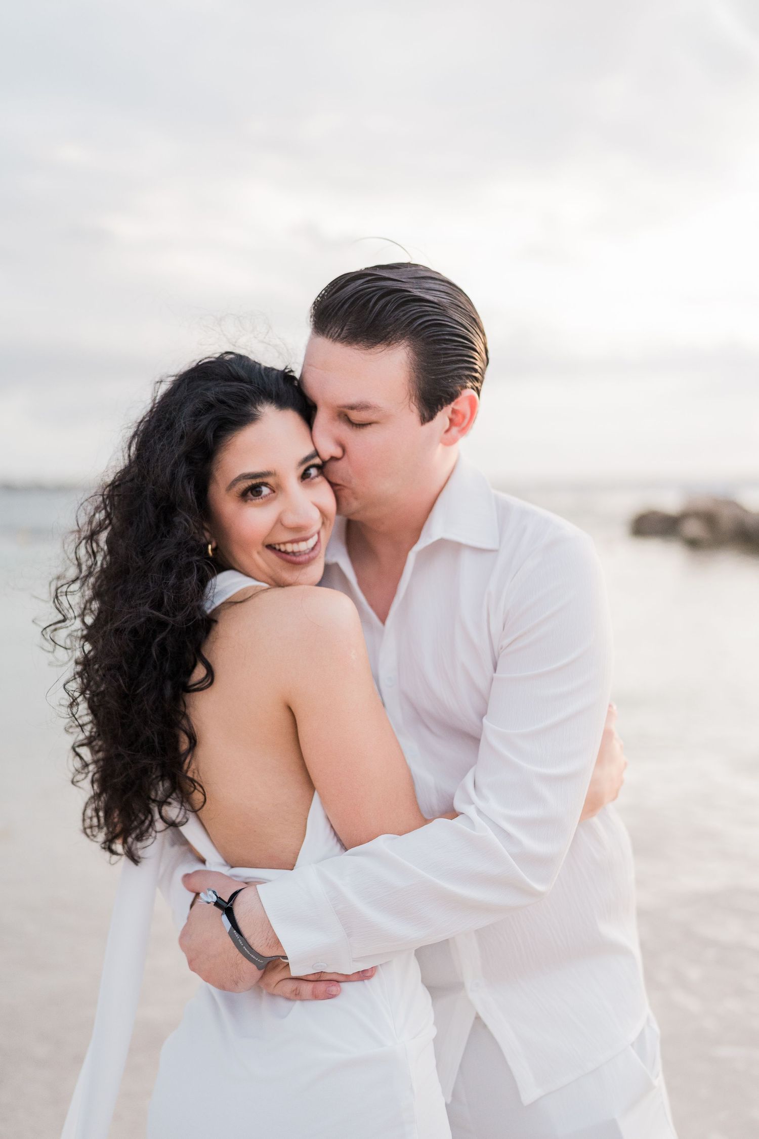Couple in white embracing on a beach, woman smiling as man kisses her cheek, with ocean backdrop.