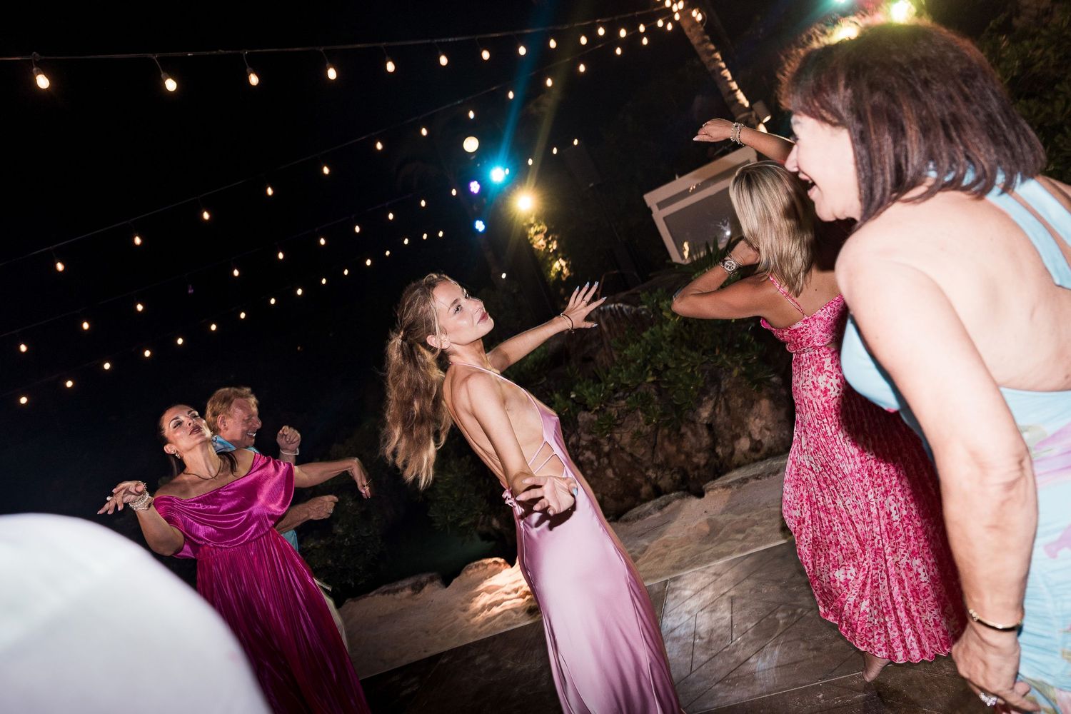 Women in pink and sequin dresses dancing outdoors at night under string lights at an elegant party.