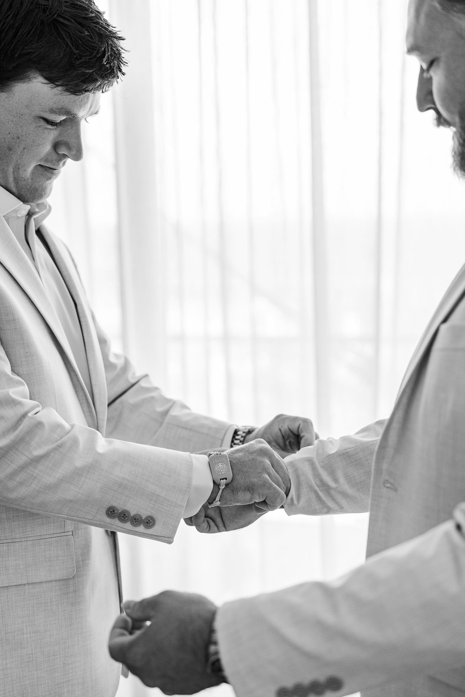 Black and white photo of two men in light suits, one helping the other fasten a watch or cufflink before a wedding.