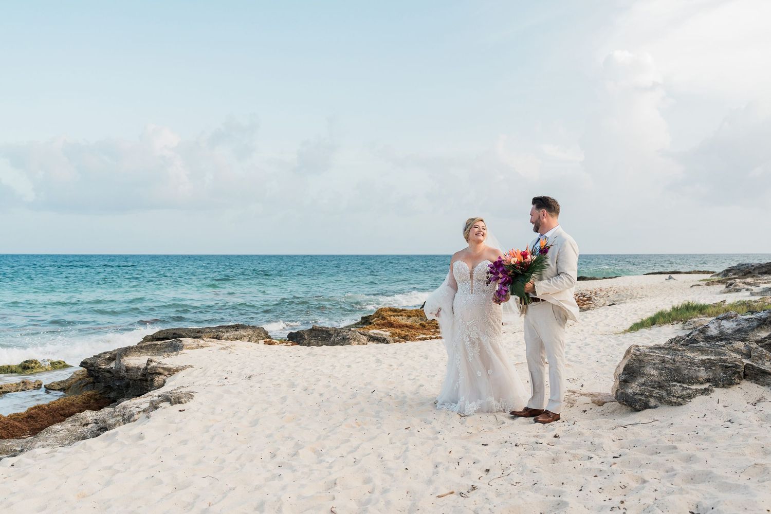 Hotel Xcaret México Wedding -Bride and groom stand on a white sandy beach holding a colorful bouquet with turquoise ocean in the background.