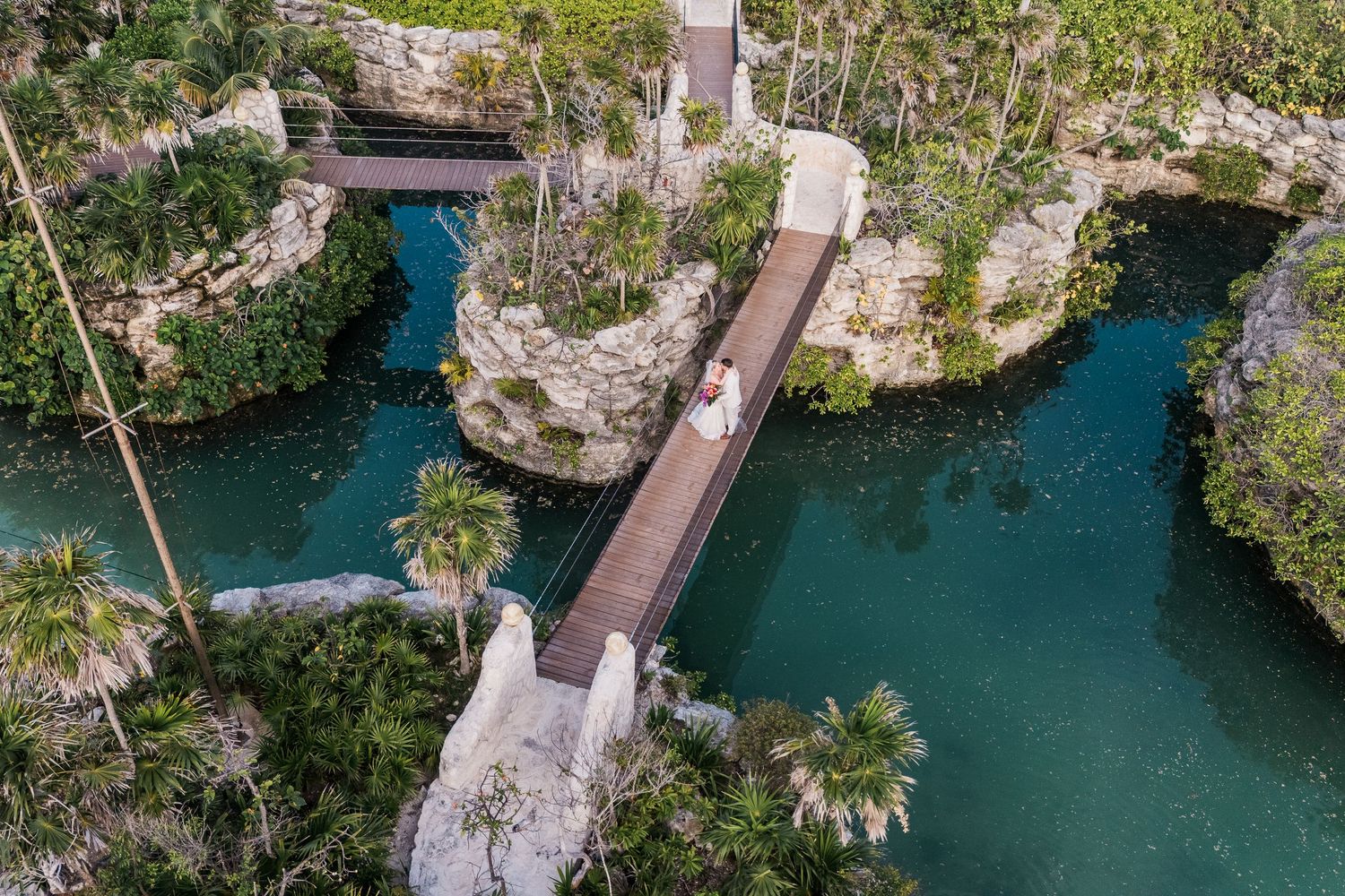 Aerial view of a bride crossing a wooden bridge over a turquoise cenote surrounded by lush tropical jungle.