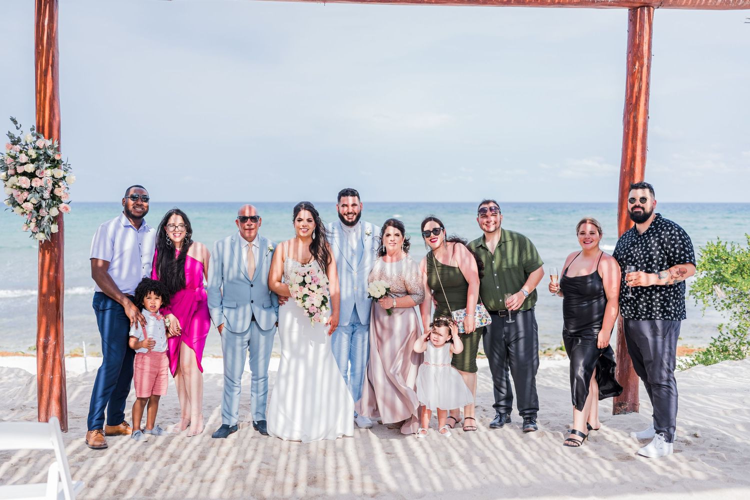 Small wedding group portrait with bride and groom surrounded by guests under a beachside pergola.