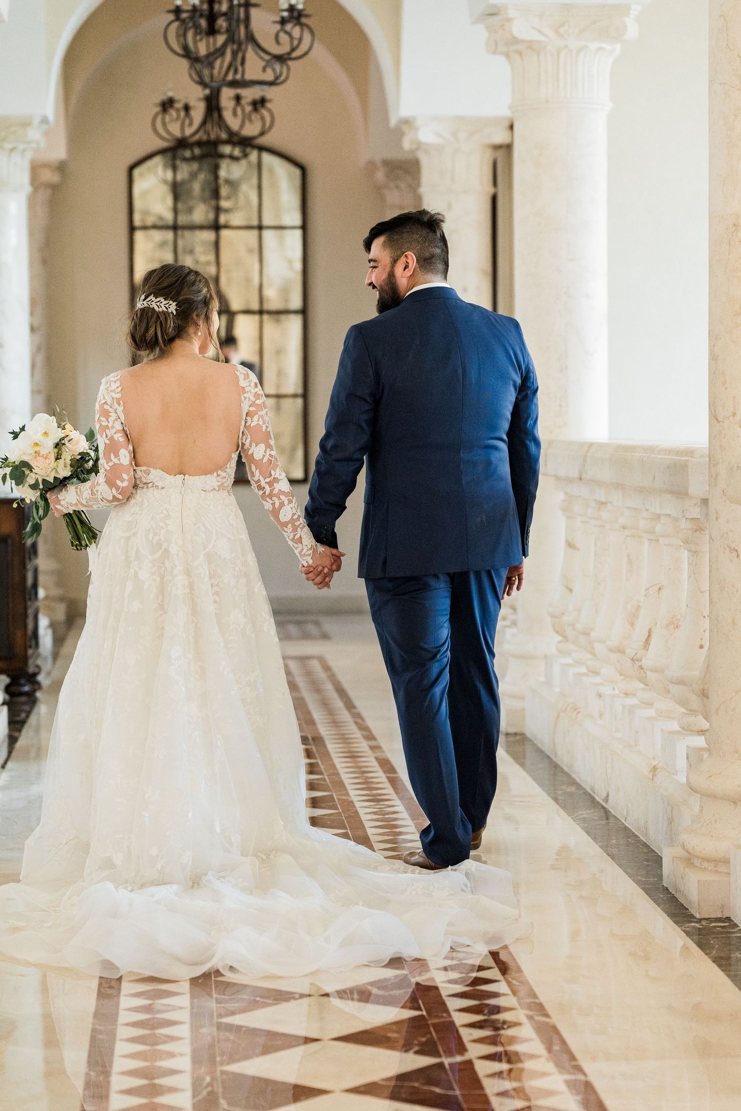 Bride and groom walk hand in hand through elegant marble hallway with tall columns.
