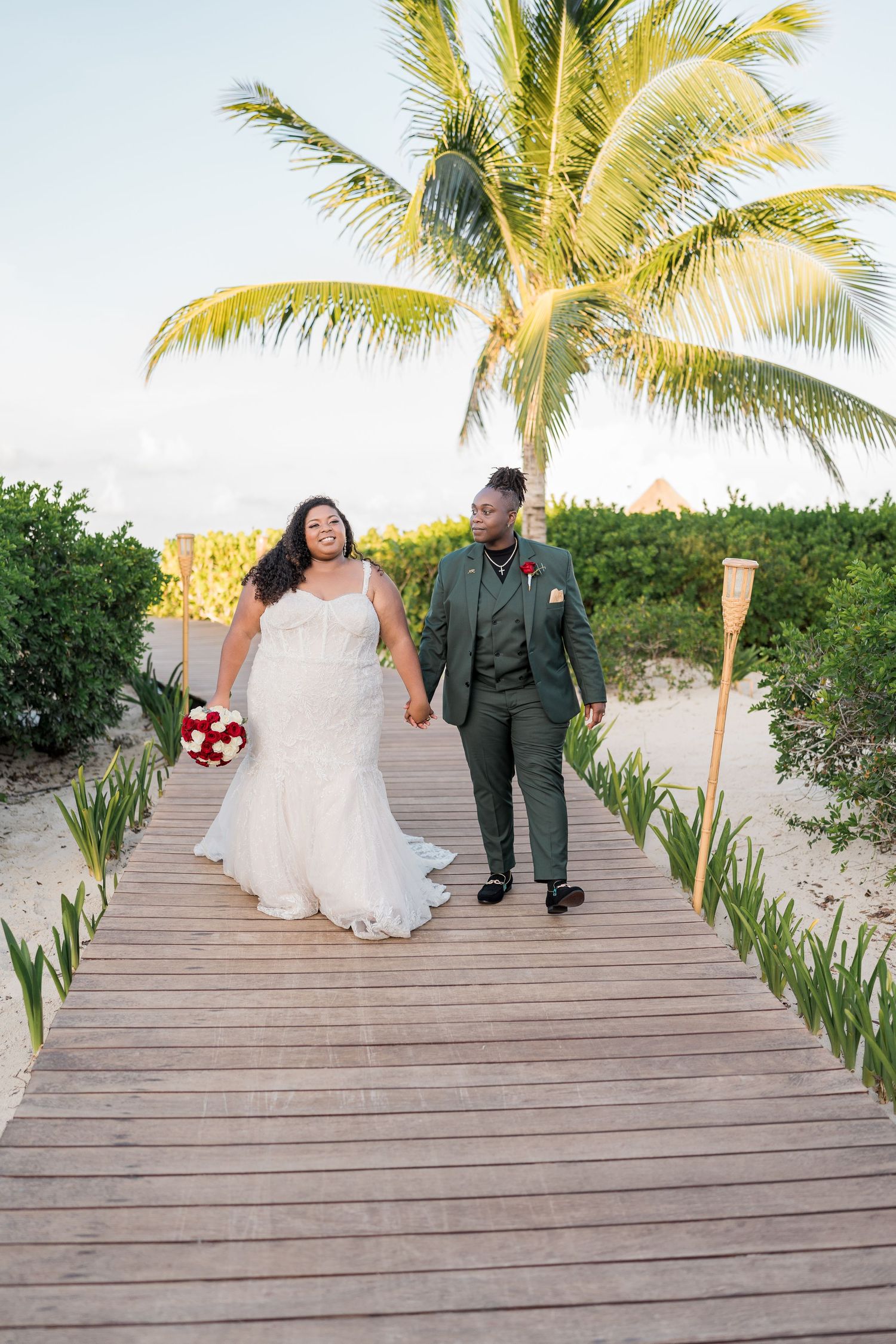 Bride and groom walk hand in hand on tropical boardwalk under palm trees.