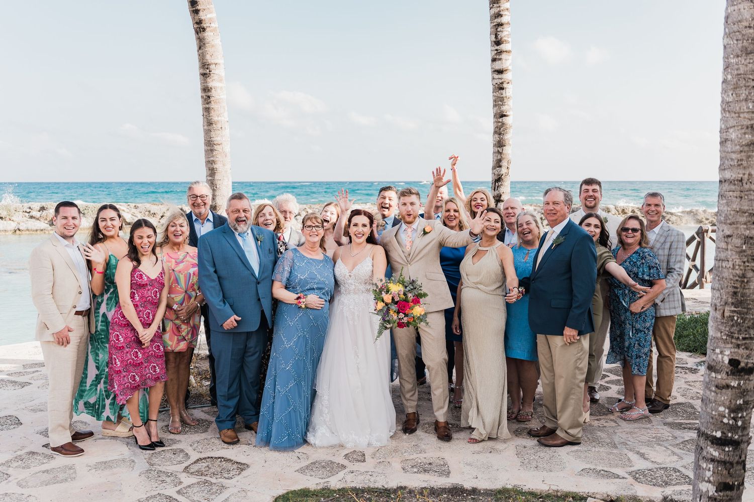 Wedding party and family pose for group photo by palm trees with ocean views at tropical venue.