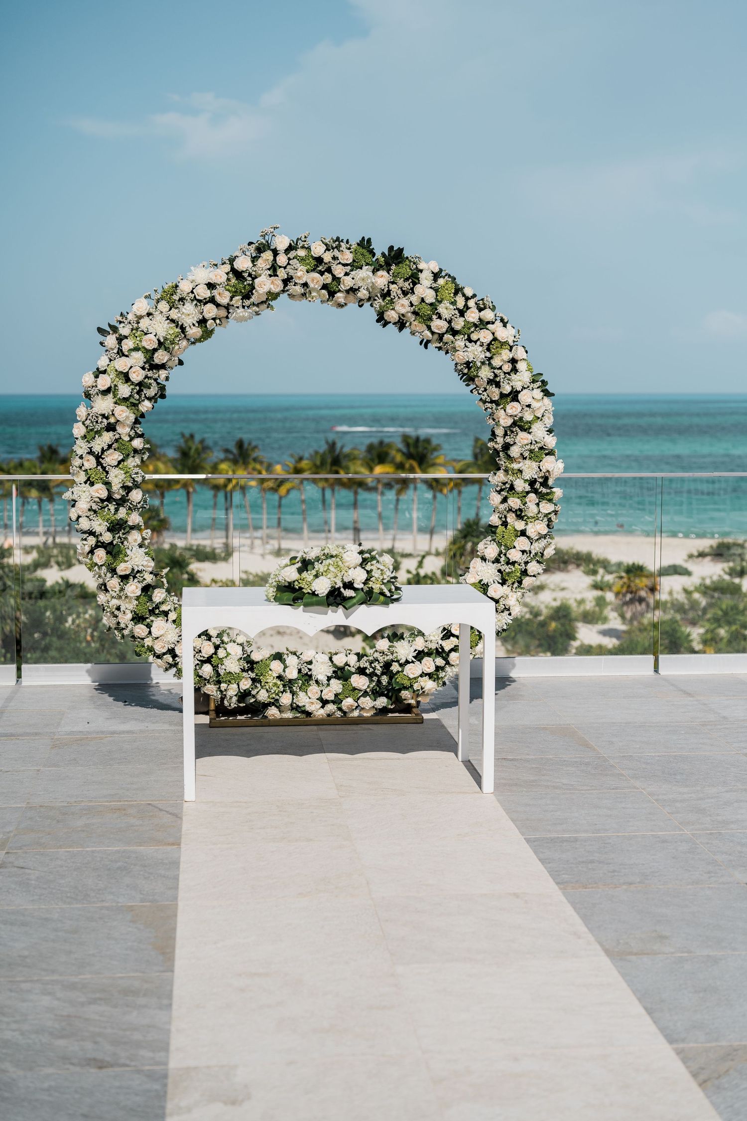 Circular white floral wedding arch adorned with roses overlooks tropical beach and turquoise sea.