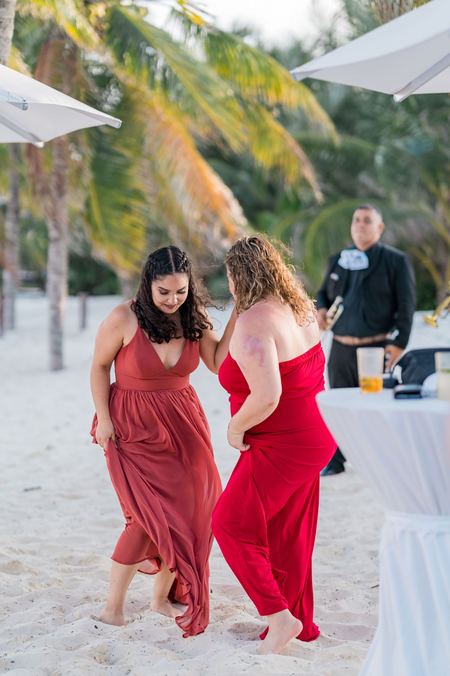Two women in red dresses dance barefoot on sandy beach during an outdoor tropical wedding reception.