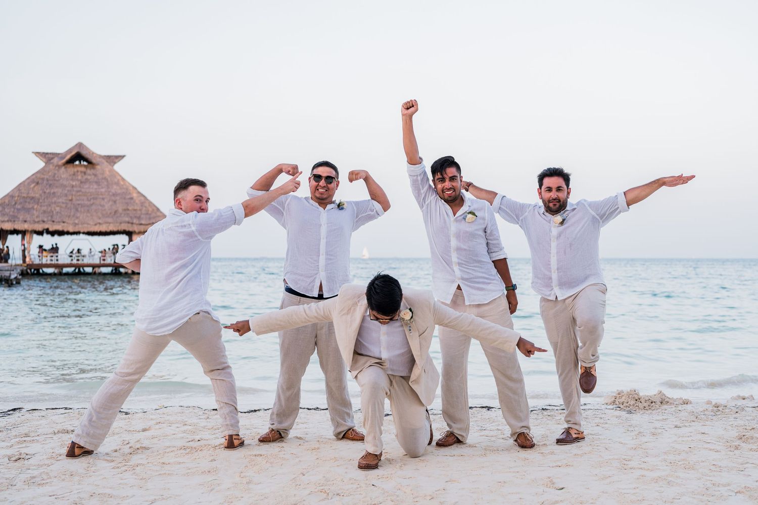 Groomsmen celebrate in white shirts and tan pants on a beach at a destination wedding.
