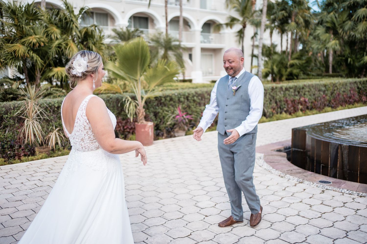 Groom reacts with joy as bride in white gown approaches during first look at tropical resort.