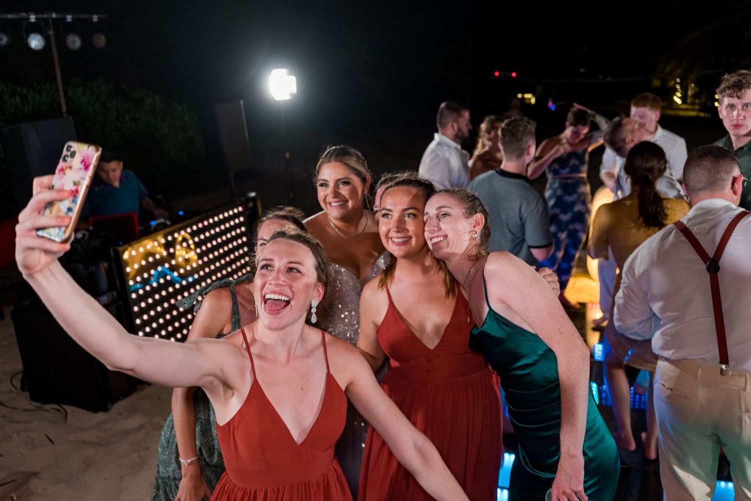 Bridesmaids in rust dresses taking a selfie at outdoor beach wedding reception.