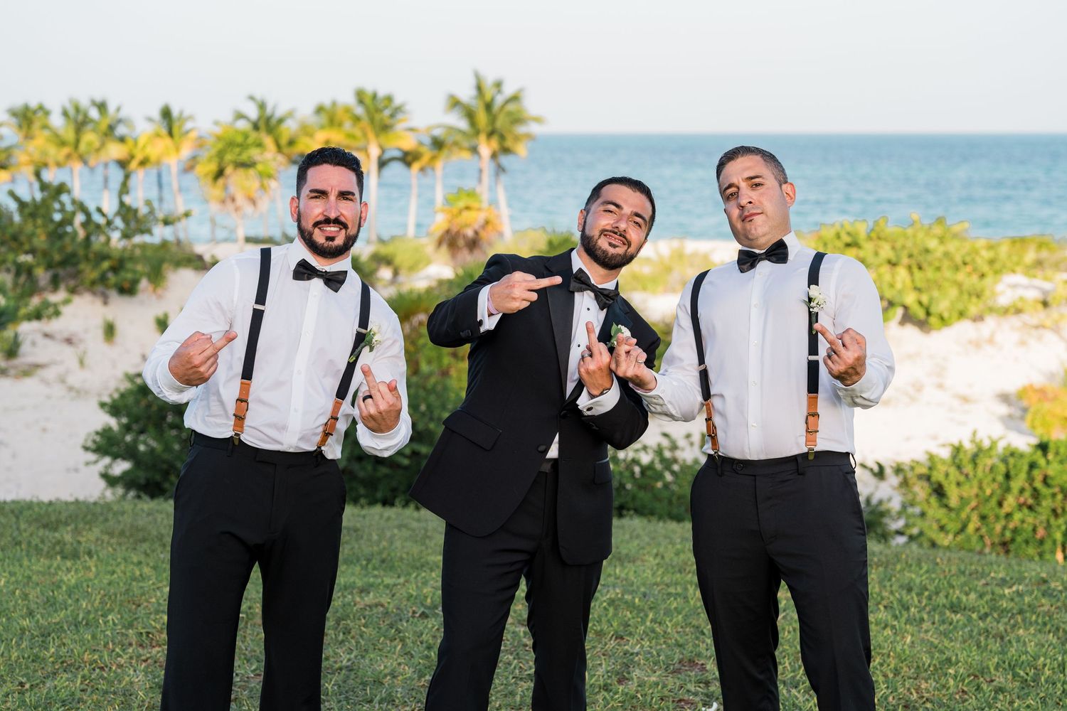 Three groomsmen in black tuxedos and suspenders smile and point at camera by the ocean.