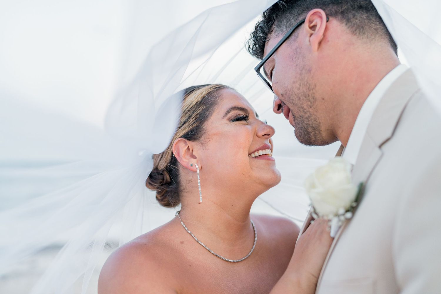 Happy bride and groom gazing at each other under a white veil, bride wearing elegant drop earrings.