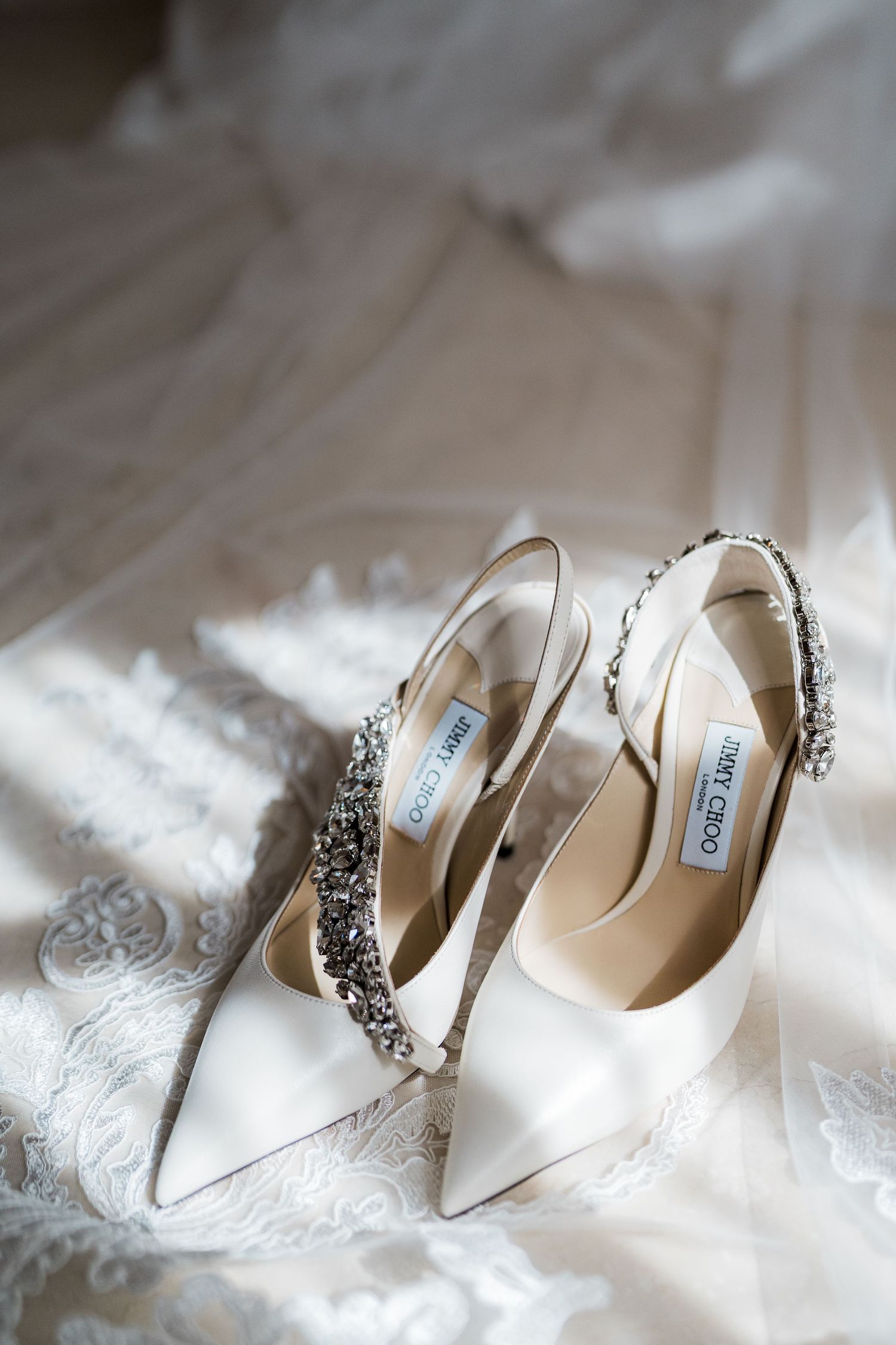 Elegant Jimmy Choo bridal heels with crystal embellishments displayed on delicate white lace wedding veil in soft natural light.