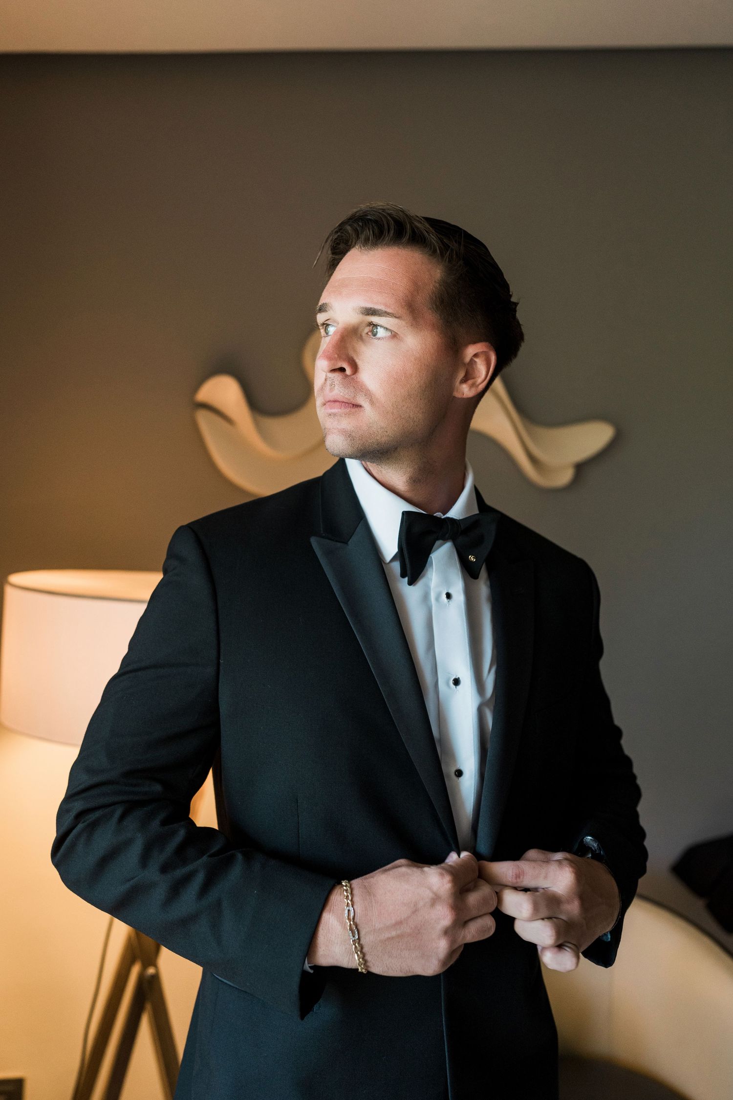 A dapper groom in a black tuxedo and bow tie buttoning his jacket while gazing away in an elegant room.