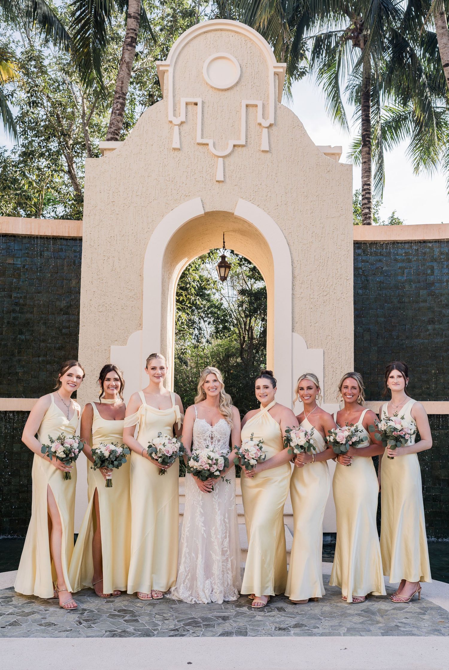 Bride in lace gown poses with seven bridesmaids in yellow satin dresses holding bouquets before an arched venue backdrop.