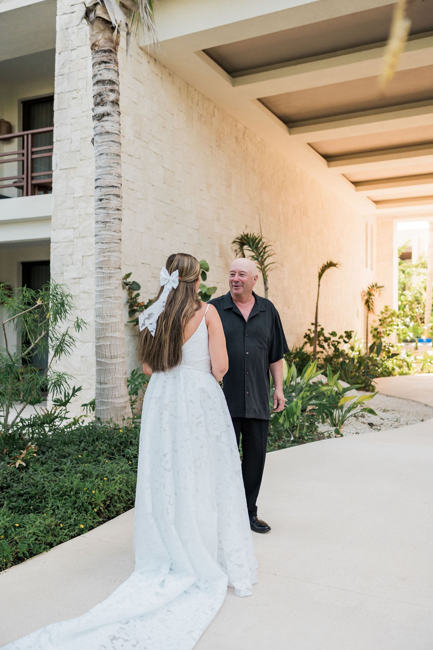Bride in white lace gown with bow shares first look moment with older man in black shirt at tropical resort.