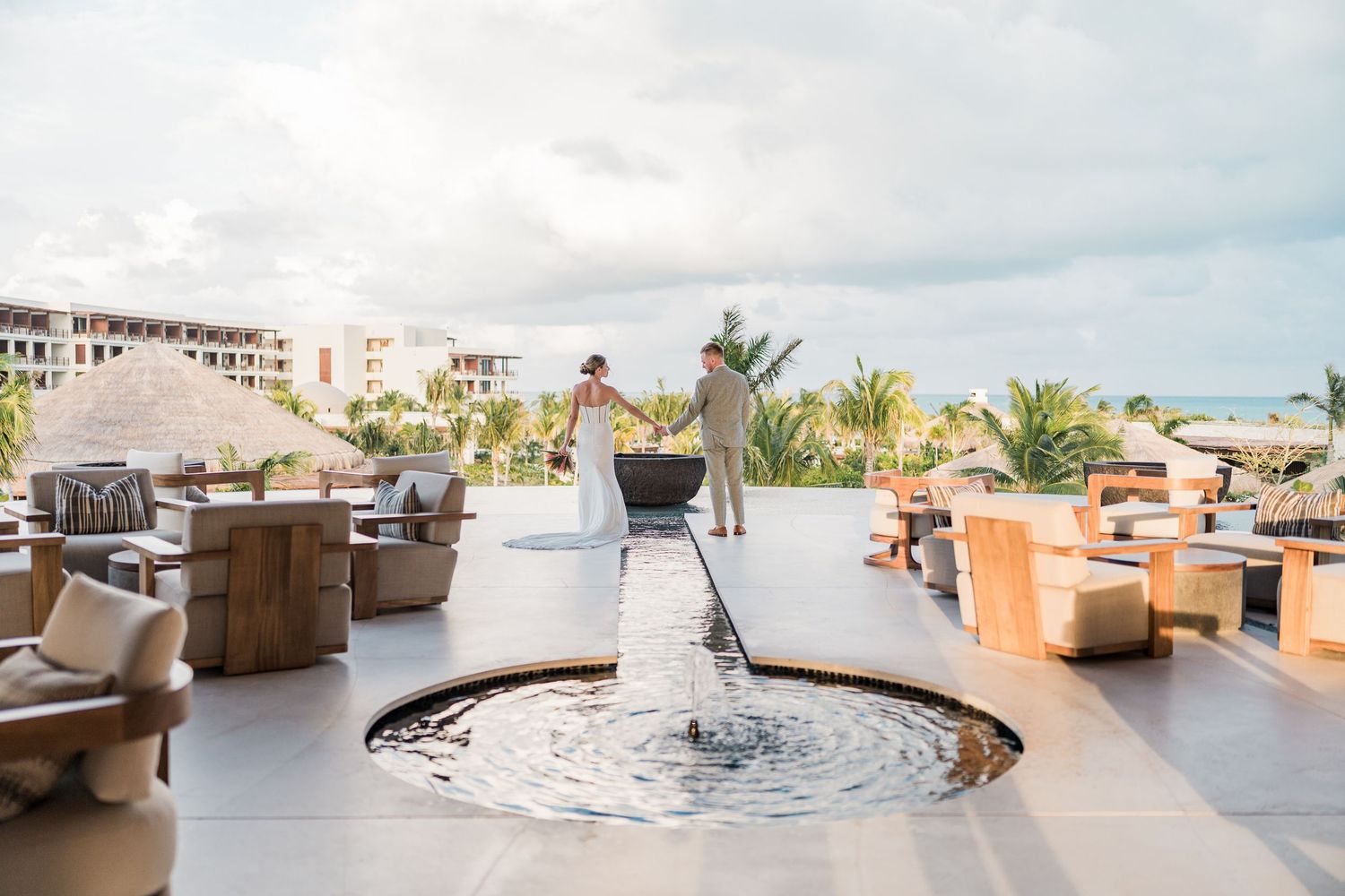 Couple standing by a circular fountain on a luxury resort terrace with ocean views and tropical landscaping.