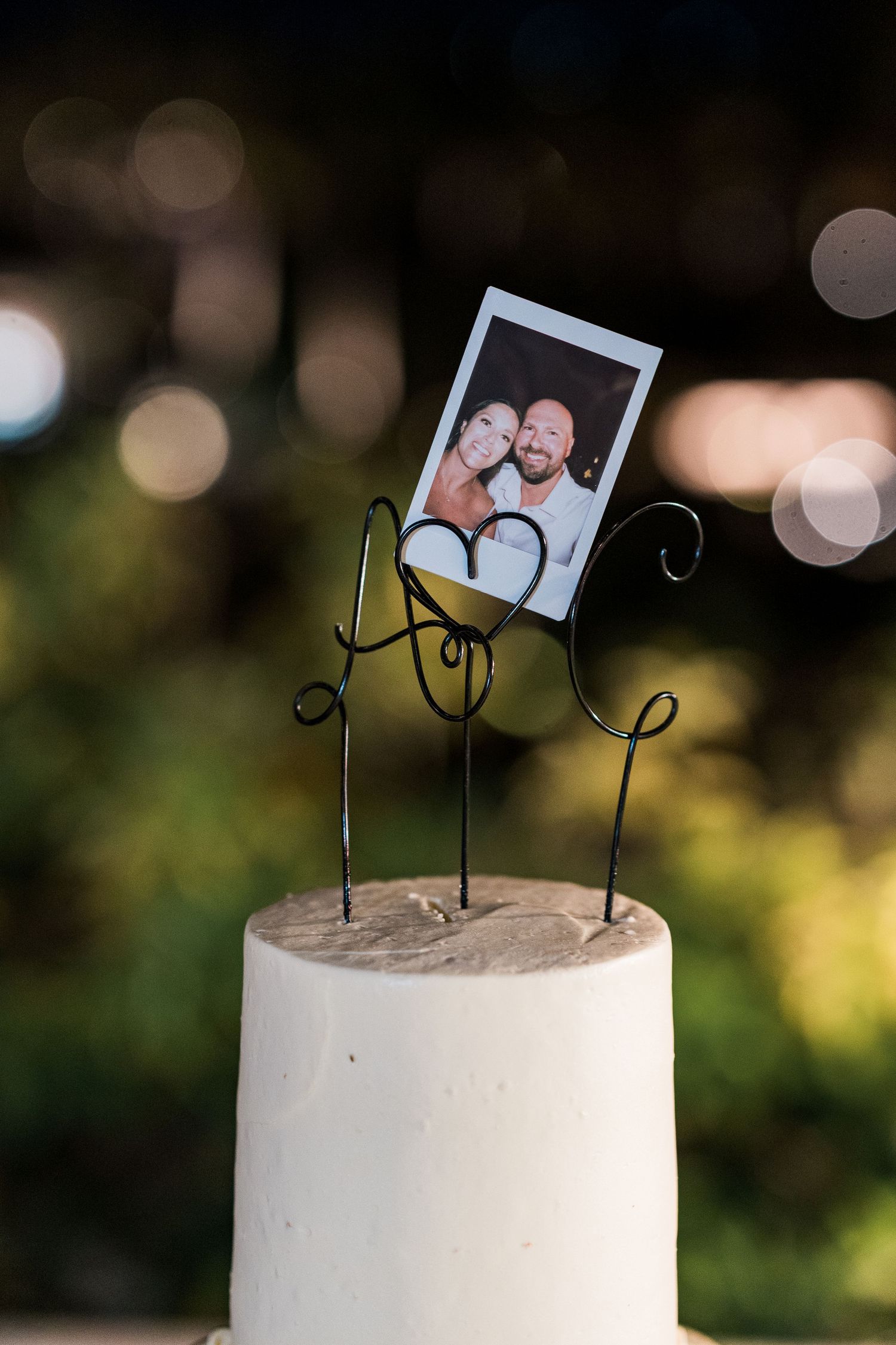 White wedding cake topped with wire cake topper holding a Polaroid photo of the happy couple, set against bokeh background.