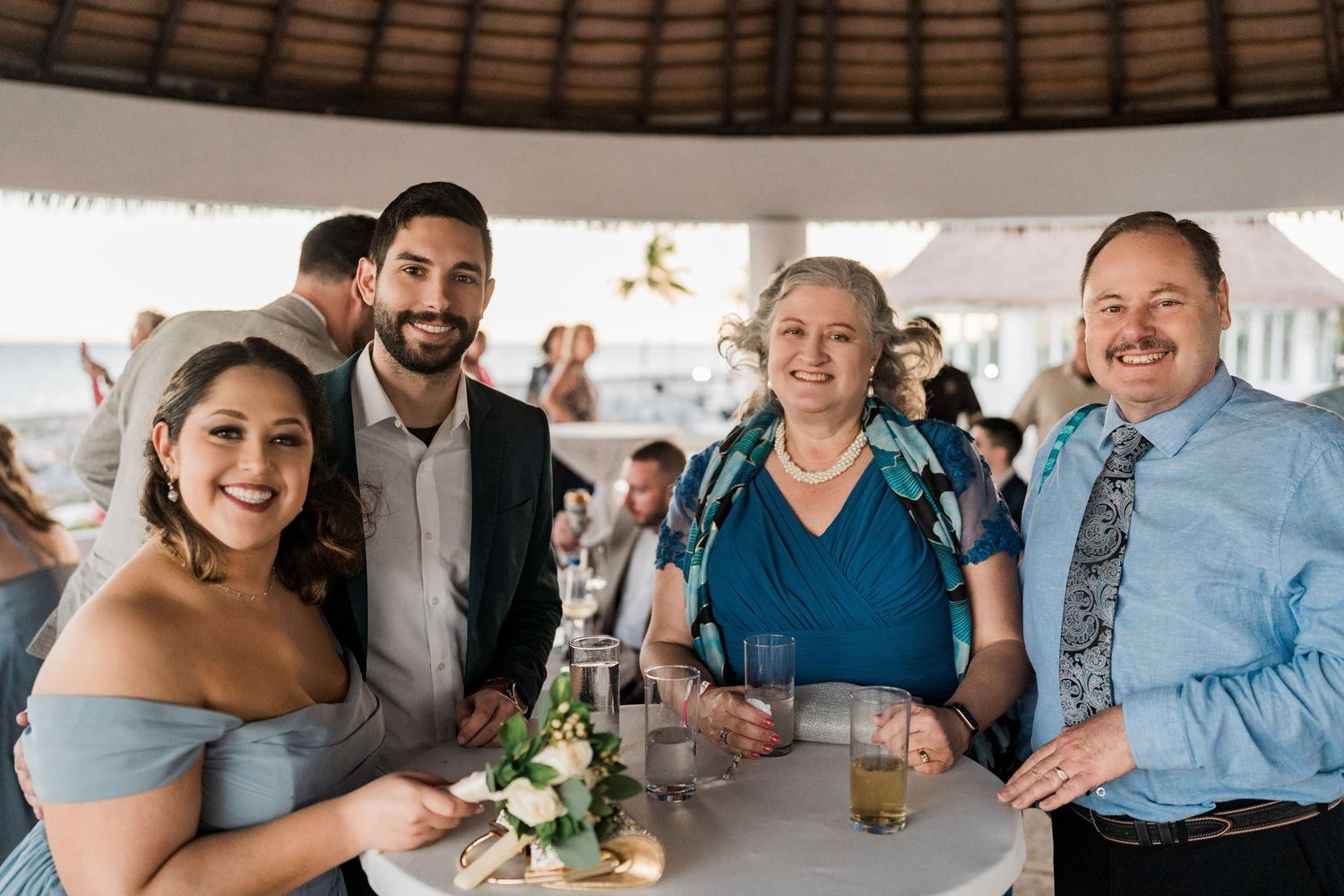 Four guests smiling together at a beach wedding reception, dressed in formal attire at a white table.