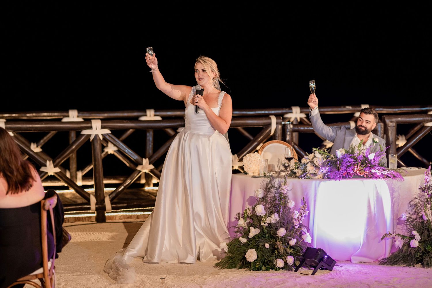 Bride in white gown raises glass during outdoor wedding reception toast at night with groom seated at floral decorated table.