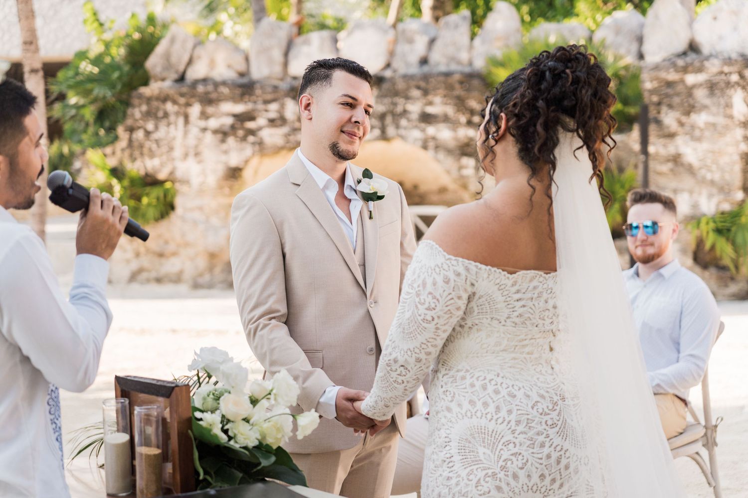 Xcaret Arte Wedding - Groom in beige suit smiles at bride in white lace dress during outdoor wedding ceremony.