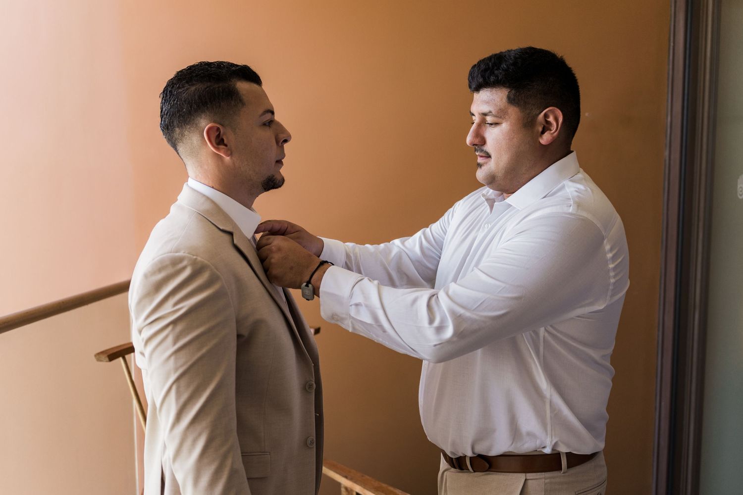 Xcaret Arte Wedding - A man in a white shirt adjusts the tie of a groom in a beige suit before a wedding ceremony.