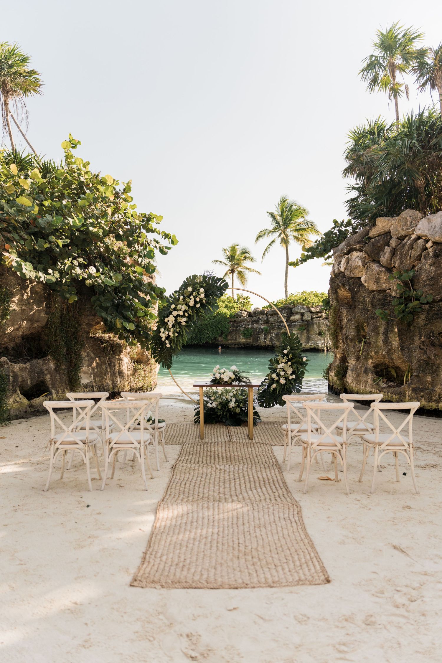 Outdoor beach wedding setup with circular floral arch, white chairs, and woven aisle runner framed by tropical rocks and turquoise water.