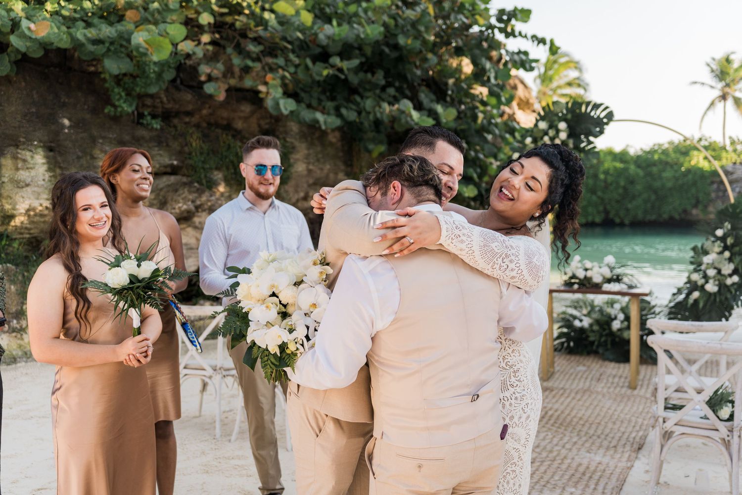 Xcaret Arte Wedding - Bride and groom share a joyful hug with guests at a beach wedding ceremony with tropical greenery.