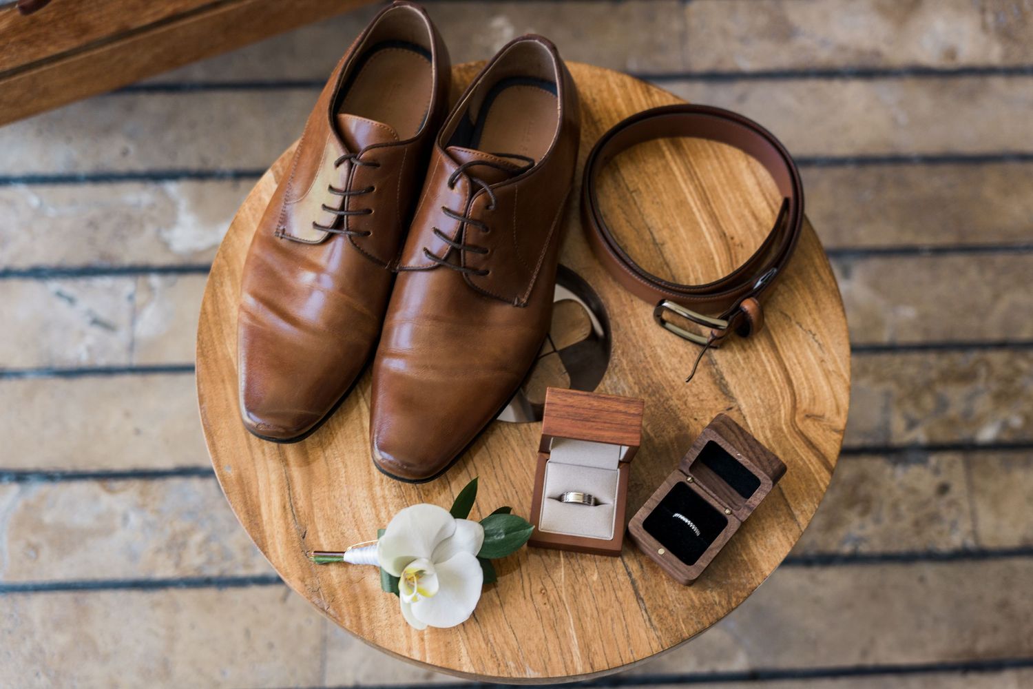 Xcaret Arte Wedding - Brown leather dress shoes, belt, ring box, wedding rings, and boutonniere arranged on a wooden table for a groom.